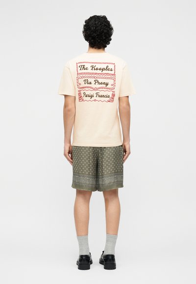 Άνδρας που φοράει μπεζ μπλουζάκι με κόκκινα γράμματα στην πλάτη και ελιάς σορτς με σχέδιο, γκρι κάλτσες και μαύρα παπούτσια, στέκεται γυρισμένος προς τα πίσω.