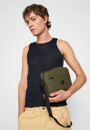 Olivegrünes rechteckiges Täschchen aus glattem Material, mit einer Vordertasche und schwarzem Zubehör. Wird von einer Person in einem marineblauen Strick-Top getragen.