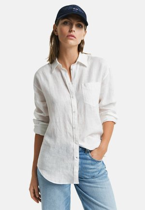 Witte linnen overhemd met een knoopsluiting aan de voorkant en een borstzak, gecombineerd met lichtblauwe denim jeans. Model draagt een donkere baseballcap.