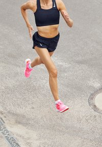 Černá sportovní podprsenka a šortky s perforovaným designem. Jasně růžové běžecké boty s bílými podrážkami a černými detaily. Běh na zpevněném povrchu.