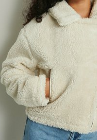Chaqueta de forro polar esponjosa de color crema con cuello y bolsillos laterales. Presenta un corte relajado y puños enrollados, mostrando una textura suave.