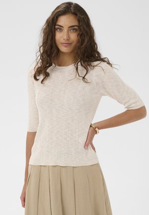 Jeune femme aux cheveux bouclés bruns, portant un haut côtelé beige clair et une jupe ample couleur sable, posant avec une main sur la hanche.