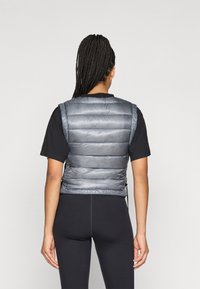 Pilka pūkinė liemenė su siūlėmis, lengvas audinys ir šiek tiek blizganti tekstūra. Sukurta su aukštu apykakle ir šoninėmis reguliavimo detalėmis.