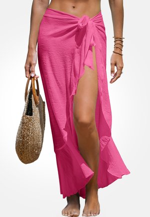 Roze lichte maxi-jurk met een gestrikt taille, met een zijopening en een gerimpelde zoom. Model houdt een geweven tas vast, blootsvoets op een lichte achtergrond.