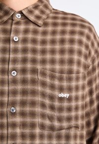 Brown and beige plaid button-up shirt with a pocket, featuring a white "obey" logo stitched on the left side. Smooth texture, classic collar.