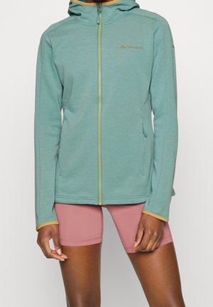 Personne portant une veste à capuche zippée turquoise avec des bordures jaunes et un short ajusté rose, debout devant un fond uni.