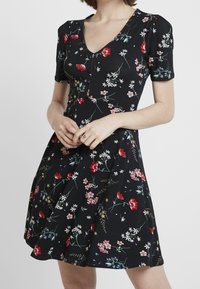 Černé květinové šaty s krátkými rukávy, výstřihem do V a knoflíkovým detailem. Mají různé barevné květiny na texturované tkanině, která se rozšiřuje od pasu.