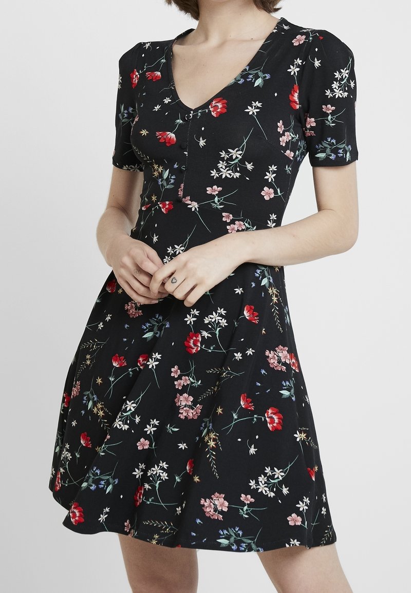 Černé květinové šaty s krátkými rukávy, výstřihem do V a knoflíkovým detailem. Mají různé barevné květiny na texturované tkanině, která se rozšiřuje od pasu.