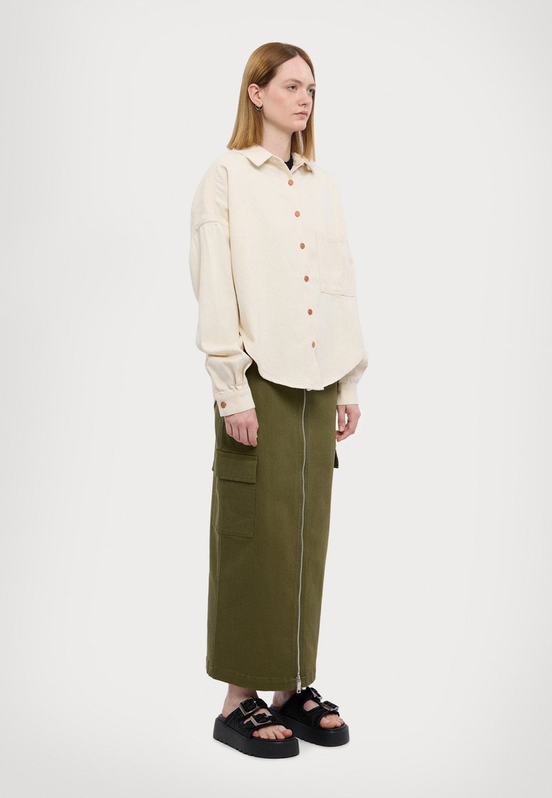 Camisa de botones color crema con un solo bolsillo en el pecho, combinada con una falda verde oliva que presenta un zipper frontal y bolsillos laterales. Sandalias de plataforma negras.