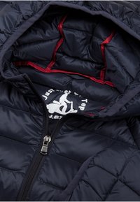 Interno della giacca a vento navy che mostra il motivo trapuntato, foderato di rosso e con zip nera. Include un logo bianco all'interno.