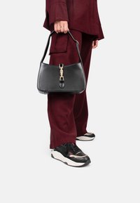 Borsa a tracolla nera in pelle con forma strutturata, dettagli in metallo dorato e manico corto, tenuta da una persona in abbigliamento bordeaux e sneakers nere.