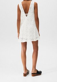 Valkoinen pitsimekko, jossa on syvä V-mallinen selkä, säädettävä nauhakiinnitys ja kerroksellinen helma. Yhdistetty mustiin puukengiin, joissa on puupohja ja niittejä.