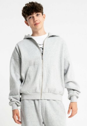 Jasnoszara bluza z kapturem na zamek, wykonana z miękkiego materiału, z kapturem, kieszeniami z przodu i elastycznymi mankietami. W połączeniu z pasującymi szarymi spodniami dresowymi.