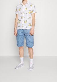 Man met een witte, knoopsluiting, korte mouwen shirt met gele citroenprint, lichtblauwe cargoshorts, witte sokken en grijze Nike sneakers.
