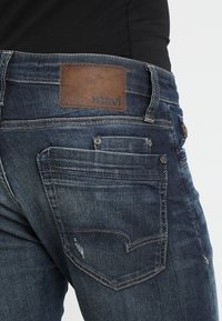 Jeans de mezclilla azul oscuro con un parche de cuero marrón que presenta la marca "mavi", costuras visibles y dos bolsillos traseros con un leve desgaste.
