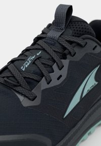 Czarne sportowe buty z teksturowaną powierzchnią, odbijającymi akcentami, szarymi sznurowadłami oraz wyraźnym logo. Posiadają wyściełany kołnierz i elastyczną podeszwę.