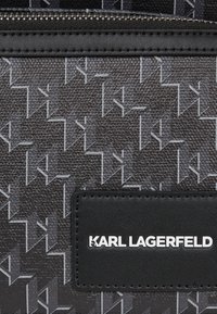 Material texturat negru cu un model geometric în gri. Prezintă o etichetă din piele neagră cu "KARL LAGERFELD" embossat în alb.