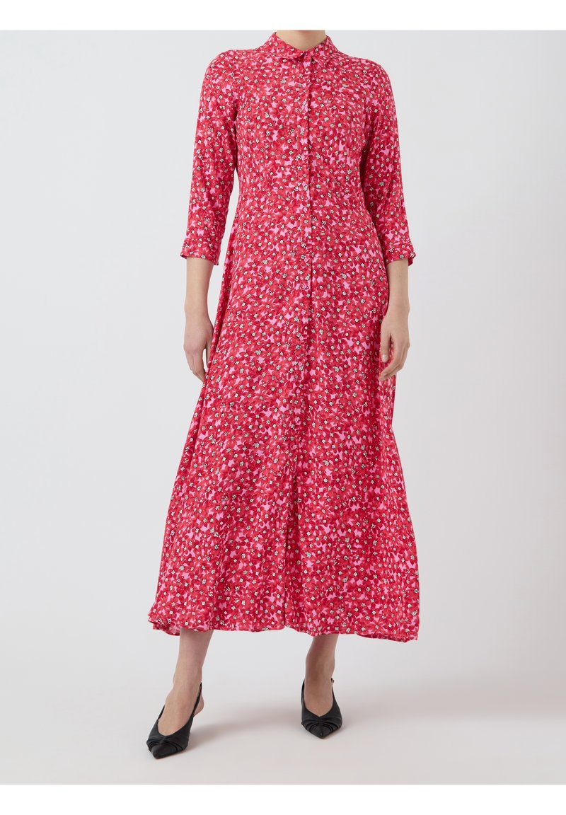 Femme portant une longue robe fleurie rouge, manches trois-quarts, boutonnée devant, et des talons noirs à bout pointu, debout sur un fond clair.