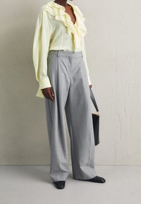 Person som bär en ljusgul blus med volanger, grå högmidjade vida byxor, svarta skor, och håller en svart handväska mot en enfärgad vägg.