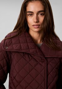 Jeune femme aux longs cheveux bruns portant une veste matelassée bordeaux avec un grand col, regardant directement la caméra.