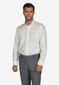 Mann in einem cremefarbenen Hemd mit Knöpfen und grauen Hosen posiert selbstbewusst vor einem neutralen Hintergrund, der sein Outfit hervorhebt.