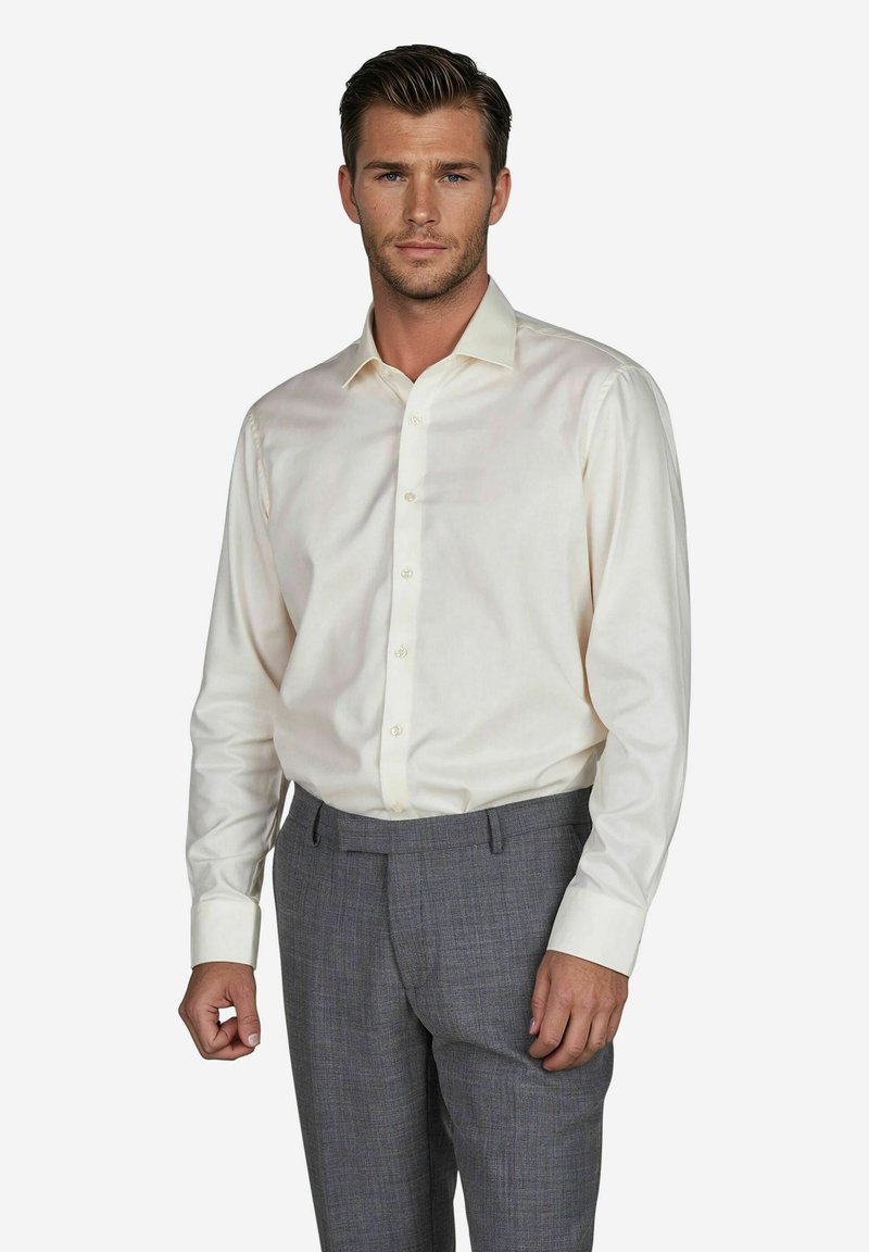 Mann in einem cremefarbenen Hemd mit Knöpfen und grauen Hosen posiert selbstbewusst vor einem neutralen Hintergrund, der sein Outfit hervorhebt.