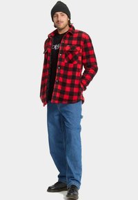 Camisa de franela a cuadros rojos y negros, diseño con botones, dos bolsillos en el pecho, combinada con jeans oscuros y zapatos negros, rematada con un gorro negro.