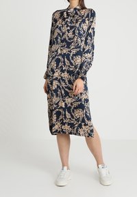 Mörkblå klänning med blommönster i beige; långa ärmar, krage och knälång nederdel; bärs med vita sneakers.