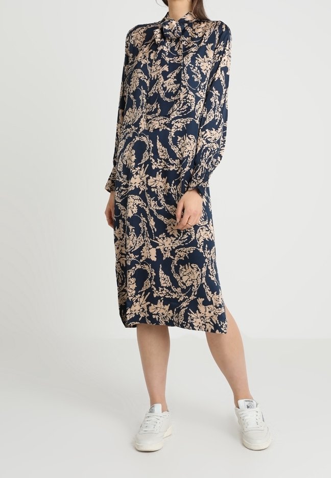 Mörkblå klänning med blommönster i beige; långa ärmar, krage och knälång nederdel; bärs med vita sneakers.