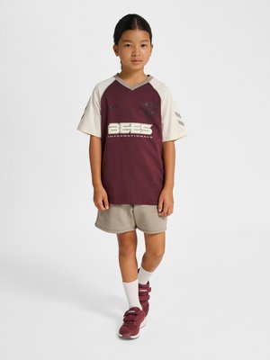 Barn med mørkt hår i flettet hestehale iført en bordeaux og beige sportstrøje, beige shorts, hvide sokker og bordeaux sneakers, står på hvid baggrund.