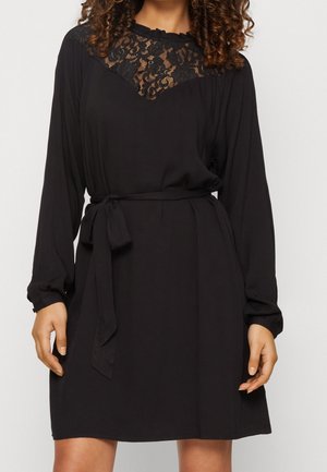 Woman wearing a black long-sleeve dress with lace detail at the neckline and a tied waist belt, standing against a plain background.