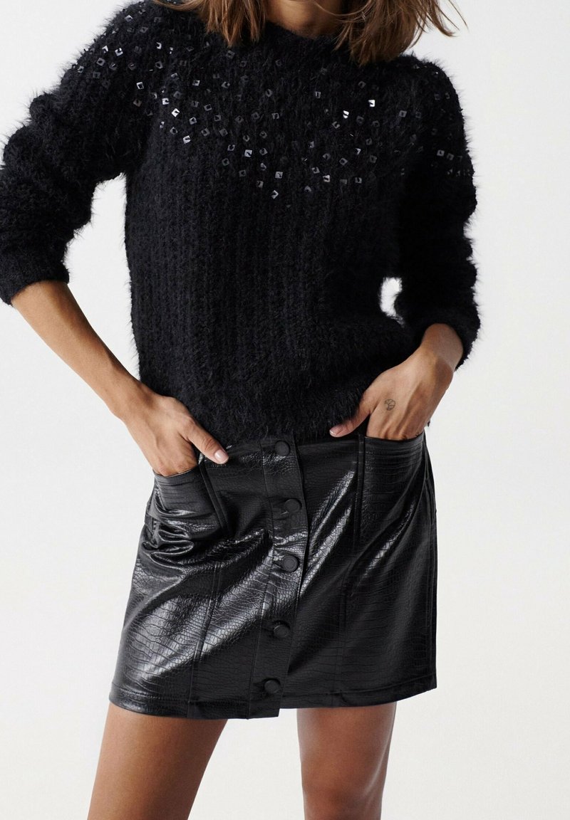 Femme portant un pull texturé noir avec des sequins et une mini-jupe noire en cuir synthétique boutonnée avec motif crocodile, mains dans les poches.