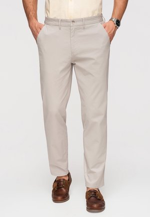 Homme portant un pantalon beige décontracté, une chemise claire, des mocassins en cuir marron et une montre-bracelet, debout les mains dans les poches.