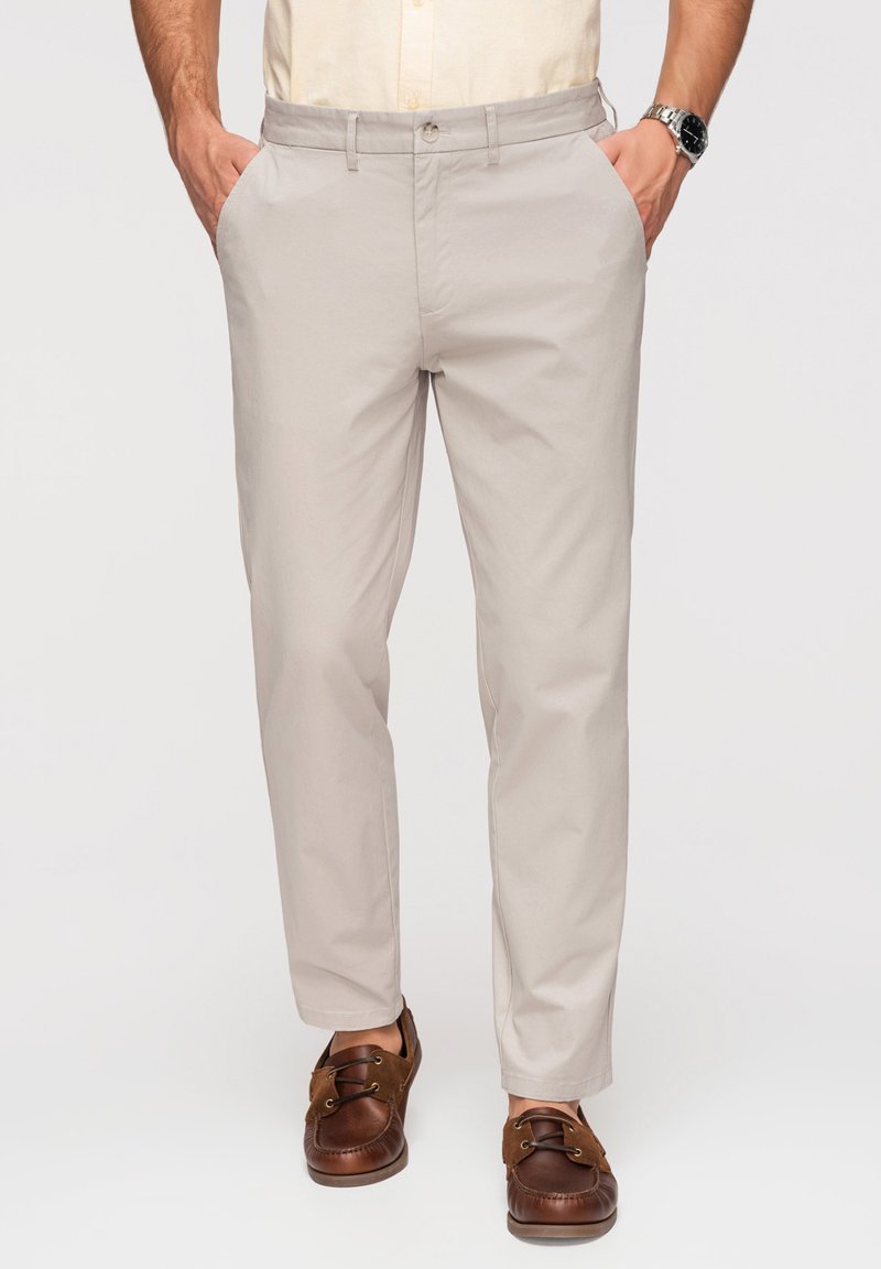 Homme portant un pantalon beige décontracté, une chemise claire, des mocassins en cuir marron et une montre-bracelet, debout les mains dans les poches.