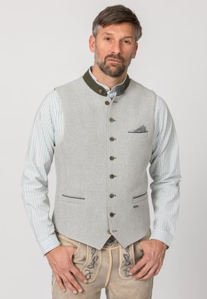 Mann trägt traditionelle beige bestickte Lederhose und hellgraue, geknöpfte Weste über gestreiftem Hemd, steht mit den Händen in den Hüften.