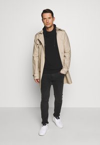 Gabardina beige con cintura ceñida, superpuesta a una sudadera negra. Jeans negros ajustados y zapatillas blancas completan el conjunto.
