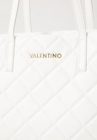 Balta pagalvėlė su auksinės metalinės „Valentino“ logotipu. Yra siuvinėta deimantų raštu ir dvi baltos rankenos. Lygus sintetinės medžiagos paviršius.