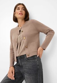 Beige knitted cardigan with golden buttons and a chest pocket, paired with a light brown blouse and gray denim jeans. Smooth texture.