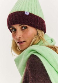 Pletená čepice v světle zelené a tmavě bordó barvě, s přehnutým lemem a malou fialovou cedulkou. Měkká textura; doplněna zeleným šátkem.