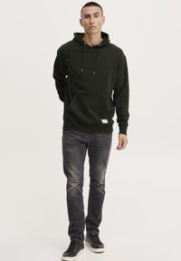 Felpa verde scuro con tasca a canguro e cordini, abbinata a jeans grigi sbiaditi e sneakers nere. Design semplice e lineare.