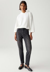 Vrouw in een witte tekstuur hoodie, vervaagde zwarte slim jeans en zwarte Mary Jane flats, staande tegen een effen lichte achtergrond.