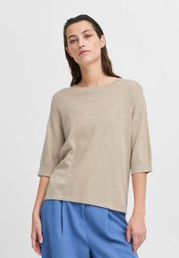 Beige lehký pletený svetr s uvolněným střihem, širokým výstřihem a krátkými rukávy. Spárován s modrými kalhotami s vysokým pasem.