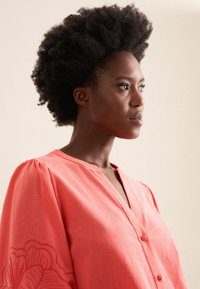 Koralowa bluzka na guziki z dekoltem w serek, bufkami i haftem kwiatowym na rękawie. Wykonana z lekkiego materiału.