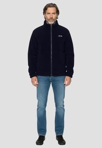 Marineblaue Fleecejacke mit hohem Kragen und durchgehendem Reißverschluss, mit strukturierter Oberfläche und Logo auf der Brust, kombiniert mit blauen Jeans und schwarzen Schuhen.