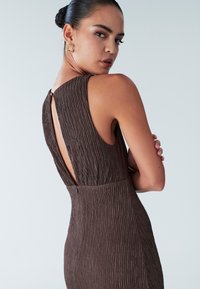 Rjava obleka z teksturo, z zaobljenim zadnjim delom, brez rokavov in diskretnim zadrgo v predelu pasu. Videz dopolnjujejo zlati uhani.