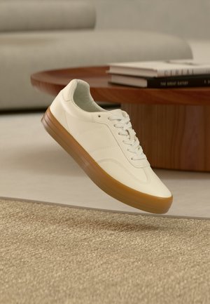 Biały tenis z gumową podeszwą unoszący się nad beżowym dywanem w salonie z drewnianym stolikiem i ułożonymi stosami książek w tle.