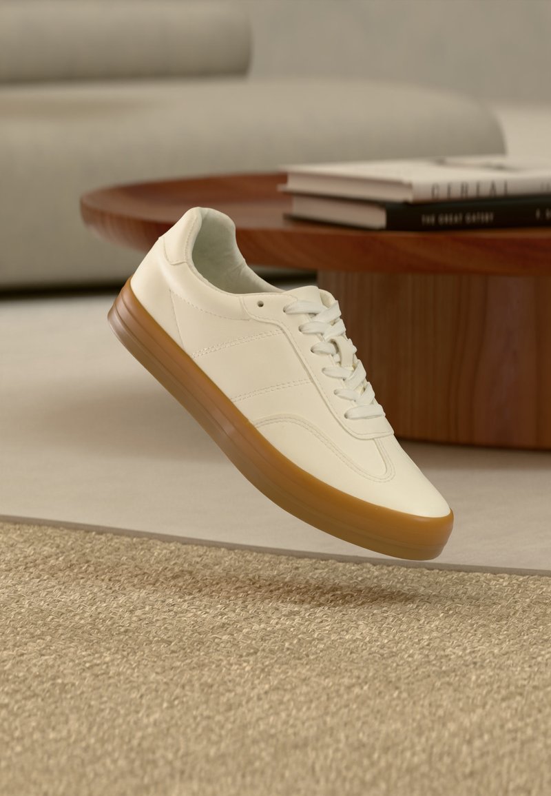 Λευκό αθλητικό παπούτσι με καφέ σόλα να αιωρείται πάνω από μπεζ χαλί σε σαλόνι με ξύλινο τραπέζι και στοιβαγμένα βιβλία στο φόντο.