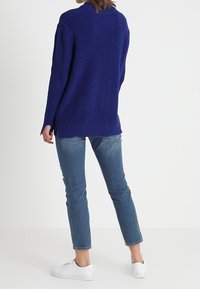 Svetr s dlouhým rukávem z vroubkované pleteniny v hluboké modré barvě, s volným střihem a bočními rozparky na lemování, v kombinaci s světlým denimem a bílými teniskami.