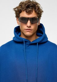 Sweat à capuche bleu avec cordon de serrage et coupe surdimensionnée ; modèle portant des lunettes de soleil angulaires, teintées et à finition brillante.