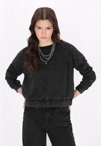 Felpa nera con finitura testurizzata, polsini e orlo a costine, abbinata a pantaloni neri coordinati. Indossa tre collane a catena in argento.